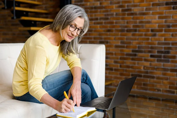 Очаровательная взрослая женщина средних лет в очках с ноутбука, сидящего на диване — стоковое фото