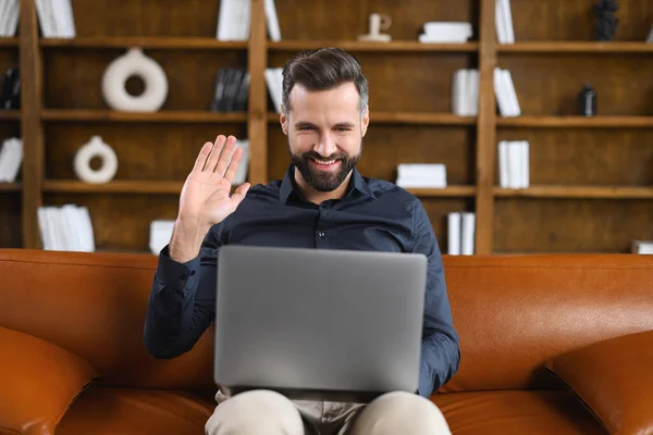 Cheerful carefree guy using laptop computer for video connection, looking at the webcam and waving hand. Man sitting at the sofa and making video call — Stock Photo, Image