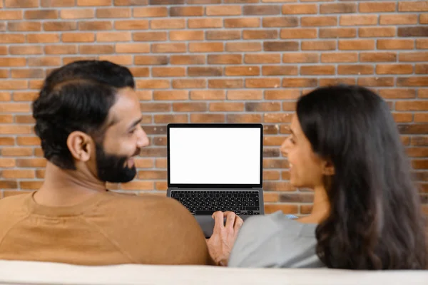 Vista posteriore della giovane coppia pakistana che utilizza il computer portatile mentre si siede sul divano a casa. I coniugi guardano qualcosa sul portatile. Felice uomo e donna rilassante — Foto Stock