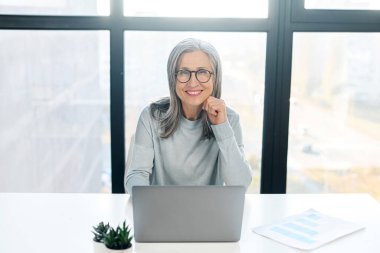 Gözlüklü, kameraya gülümseyen zarif, olgun bir iş kadınının portresi.