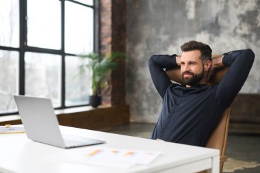 Tıraşsız bir adam, iş yerinde rahatlık ve rahatlık hissederken masada oturup gülümsüyor. Kendine güvenen bir işçi dizüstü bilgisayarın önünde oturuyor.