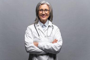 Gülümseyen gri saçlı kadın doktor kollarını kavuşturup tek başına duruyor.