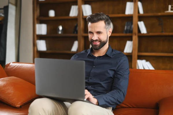Cheerful caucasian man wearing casual wear sitting on the sofa and using laptop at modern home office. Glad male entrepreneur typing on keyboard — Stock Photo, Image
