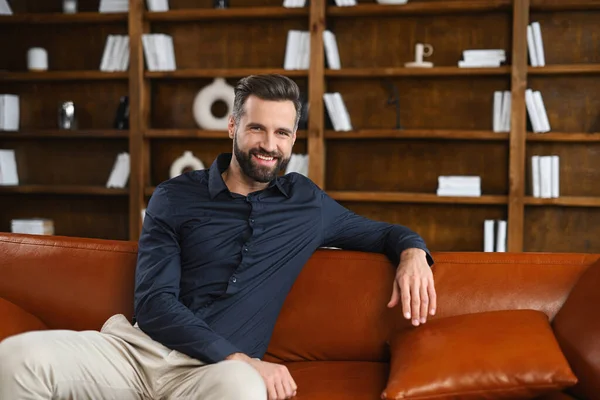 Portrait of a young caucasian man resting on sofa. Handsome positive man with hand on the back of the sofa sitting in living room, daydreaming and relaxing — Stock Photo, Image