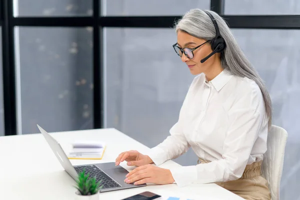 Madura mujer de negocios de pelo gris en auriculares sentados en el escritorio, reunión en línea con el cliente, videollamadas a través de computadora portátil desde la oficina. Mujer mirando la pantalla — Foto de Stock