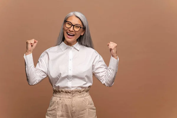 Retrato de mujer de negocios emocionada y alegre con el pelo gris de pie con los puños levantados y gritando sí, Im ganador, regocijando la victoria, el éxito. Estudio de tiro aislado en beige — Foto de Stock