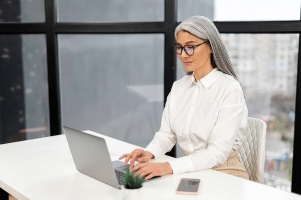 Inteligente mujer de negocios de pelo gris de edad avanzada usando una computadora portátil en la oficina. Mujer madura inteligente emprendedor mirando a través de correos electrónicos, analiza las tareas, comprobar informe —  Fotos de Stock