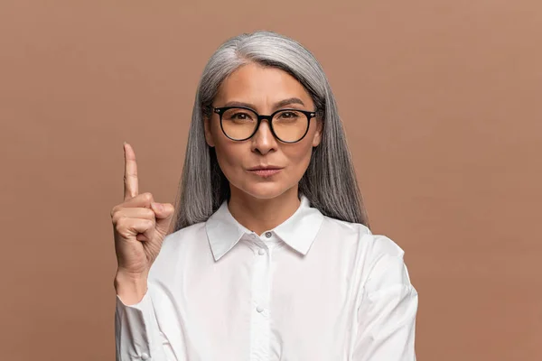 Eureka. Retrato de mujer madura asiática inspirada con el pelo largo y gris apuntando con el dedo hacia arriba con la idea de genio, habiendo inventado la solución inteligente. Estudio interior plano aislado en beige — Foto de Stock