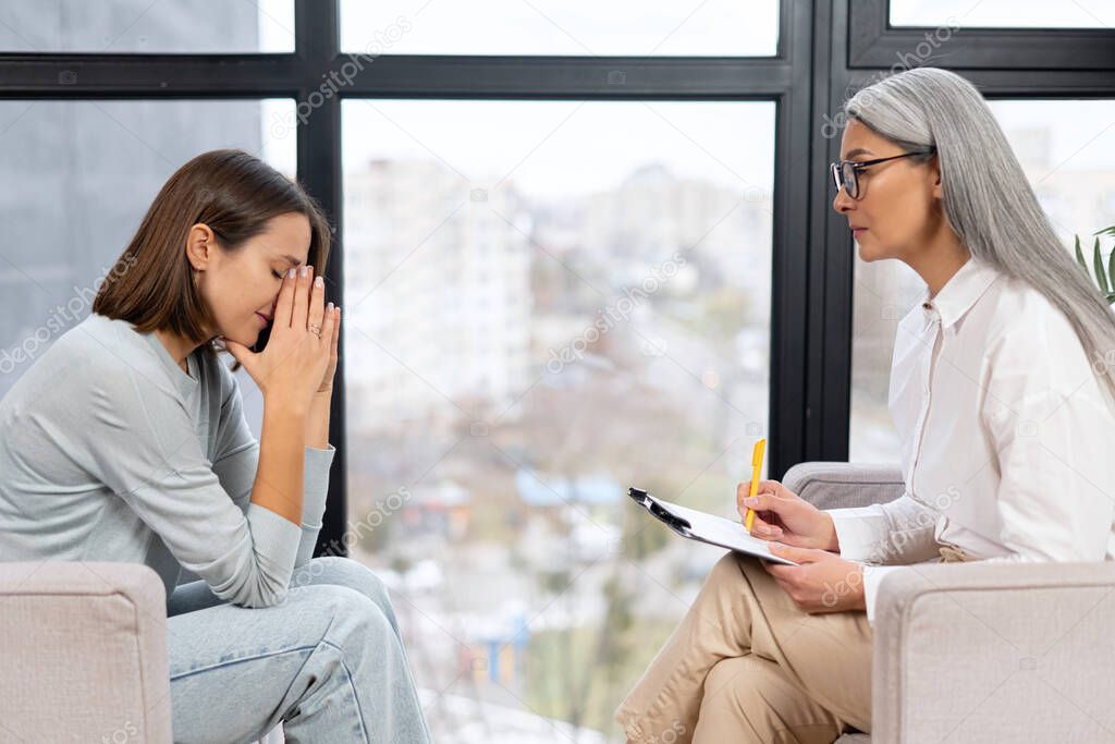 Worried young woman patient sitting at a psychologists therapist appointment and telling about mental problems, attentive mature female doctor is listening and making notes. Psychotherapy