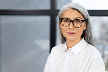 Kendinden emin ve gülümseyen yaşlı iş kadınının portre görüntüsü. Gözlüklü yaşlı kadın kameraya zevkle bakıyor. Ofis konseptinde çalışan işçi.