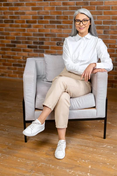 Vista completa de la mujer mayor moderna positiva y saludable sentada en el sillón en la sala de estar moderna y mirando a la cámara. Encantadora anciana soñando. Estilo de vida saludable — Foto de Stock