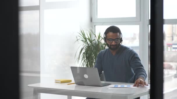 Hombre joven indio guapo con auriculares y gafas elegantes usando el ordenador portátil en la oficina — Vídeos de Stock