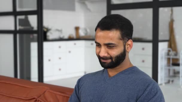 Optimistische etnische man in casual kleding met behulp van laptop zitten op de bank — Stockvideo