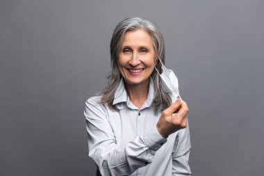 Resmi olarak giyinmiş mutlu bir yaşlı kadın. Gri üzerine izole edilmiş tıbbi maskesini çıkarıyor. Kendini viral hastalıklardan koruyan kadın ofis çalışanının yakın plan portresi. Önerilen tedbirler