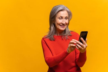 Akıllı telefon tutan neşeli, sosyal ağlarda sohbet eden, tebrik mesajları atan, gülümseyen, mobil uygulama kullanan, sarıda izole edilmiş son sınıf öğrencisi bir kadın.