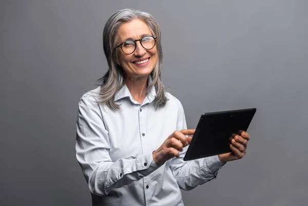 Modern elderly senior woman in formal wear using digital tablet isolated on gray. Portrait of mature female office employee using online technology for doing business — Stock Photo, Image