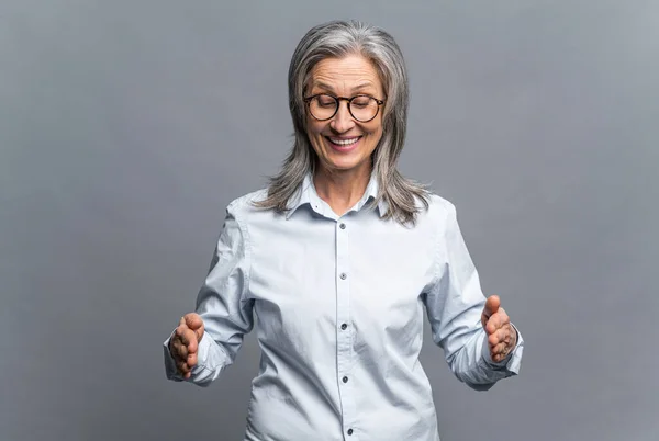 Senior madura mujer de negocios de pelo gris que muestra con las manos de longitud, medir símbolo aislado en gris. Mujer gerente en gafas de pie y demostrando el tamaño de algo grande — Foto de Stock