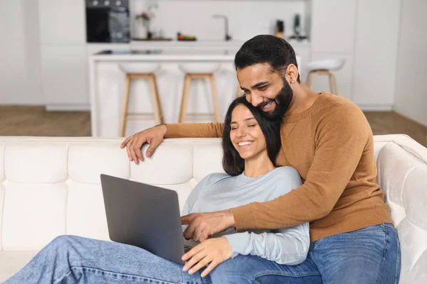 Joven pakistaní apuntando a la pantalla mientras usa el portátil en el sofá de su casa con su esposa. Esposos viendo algo en el portátil. Hombre y mujer abrazando y relajando —  Fotos de Stock