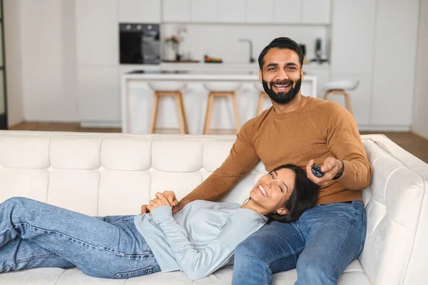 Foto horizontal de la pareja india enamorada sentada en el sofá y viendo la televisión con sonrisas de placer. Hombre multirracial sosteniendo el mando a distancia a mano —  Fotos de Stock