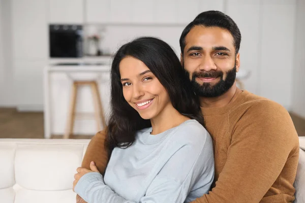 Primer plano de feliz pareja interracial posando mientras se sienta en el sofá con fondo de cocina. Felices propietarios de nuevo piso abrazando delante de la cámara —  Fotos de Stock