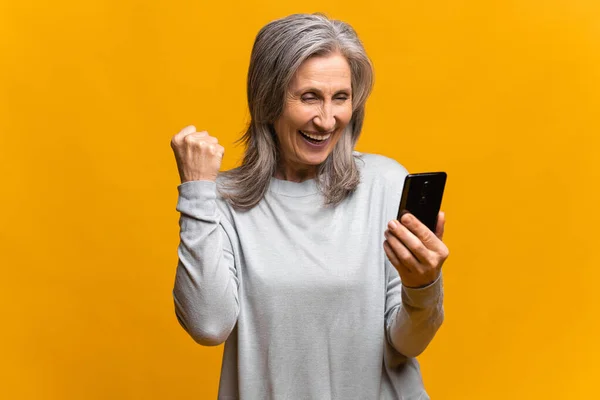 Portrait of excited overjoyed senior female shouting for joy, screaming with mobile phone in hand, celebrating online betting win, successful internet earnings. Indoor studio shot