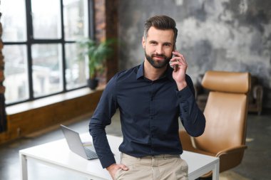 Modern bir çatı katında dururken akıllı telefondan konuşan beyaz, erkek, küçük işletme sahibi. Gülümseyen, çağdaş menajer gündelik kıyafet bekleme telefonunda.