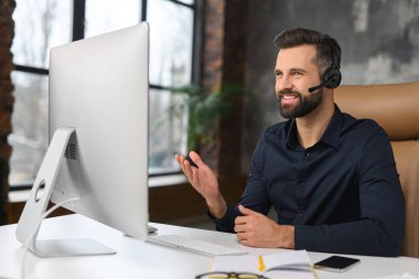 İşyerinde internetten konuşmak için kulaklık ve bilgisayar kullanan neşeli bir adam. Ofis masasında oturan ve gülümseyerek çalışan kendine güvenen bir adam.