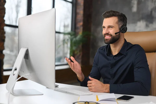 Chico alegre usando auriculares manos libres y la computadora para hablar en línea en su lugar de trabajo. Hombre seguro sentado en el escritorio de la oficina y trabajando con sonrisa — Foto de Stock