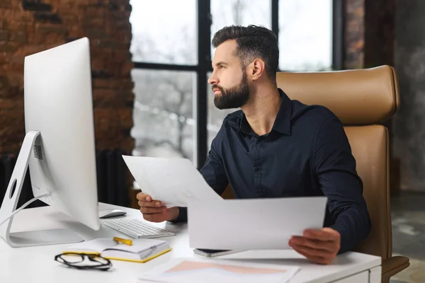 Zeki ve konsantre beyaz adam evrak işleriyle uğraşıyor. Ofisinde bilgisayarla masada oturan bir adam kağıtları karıştırıyor. Adam kontrol raporu — Stok fotoğraf