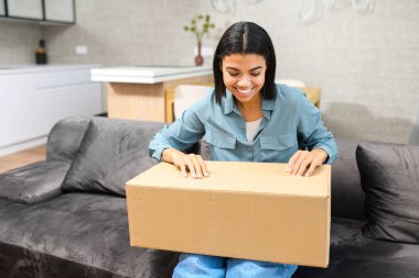 Kucağında karton kutusuyla kanepede oturan çok ırklı esmer kadın, hediye aldı, web sitesinden sipariş edilen istekleri merak ediyor.