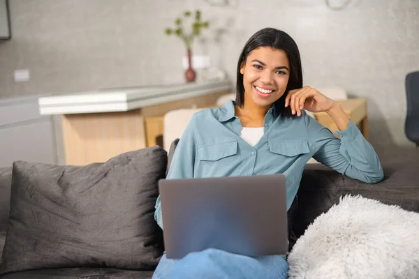 Fiatal vegyes fajú női szabadúszó barna hajjal kényelmes alkalmi ruhákban ül a kanapén és dolgozik a projekten, e-mailt ír a laptopon — Stock Fotó
