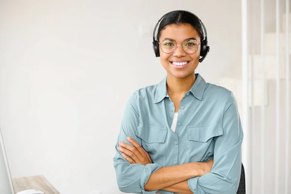 Φωτογραφία από χαμογελαστή νεαρή υπάλληλο που φορούσε ασύρματα ακουστικά, κοιτάζοντας την κάμερα. Χαρούμενη γυναίκα με ακουστικά που δέχεται κλήσεις από συναδέλφους ή πελάτες στο διαδίκτυο, που εργάζονται εξ αποστάσεως από το σπίτι — Φωτογραφία Αρχείου