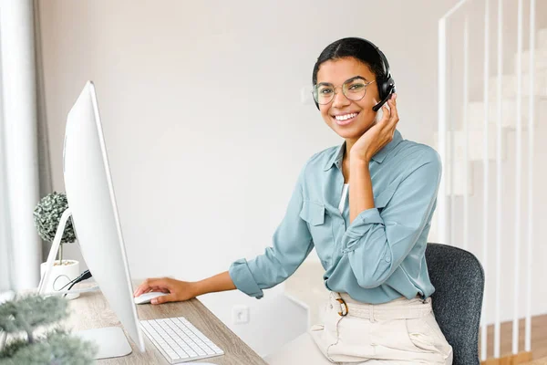 Joven mujer afroamericana que usa auriculares con computadora para el trabajo remoto mientras está sentada en la oficina del hogar. Mujer de ventas haciendo llamadas a clientes, contactos en línea —  Fotos de Stock