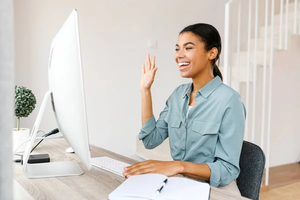 Video call. Young woman communicating via video call. She is sitting at table and waving, while looks at the screen with smile — Foto Stock