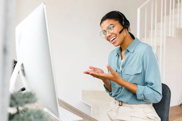 Весела приваблива жінка в бездротовій гарнітурі використовує ноутбук для відеозв'язку, розмовляючи онлайн. Продавець дзвонить, працюючи віддалено з дому — стокове фото