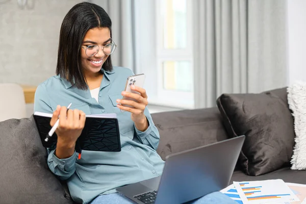 Okos afro-amerikai női szabadúszó szemüvegben ül a kanapén laptoppal az ölében, otthonról tanul és az okostelefonja képernyőjét nézi. — Stock Fotó