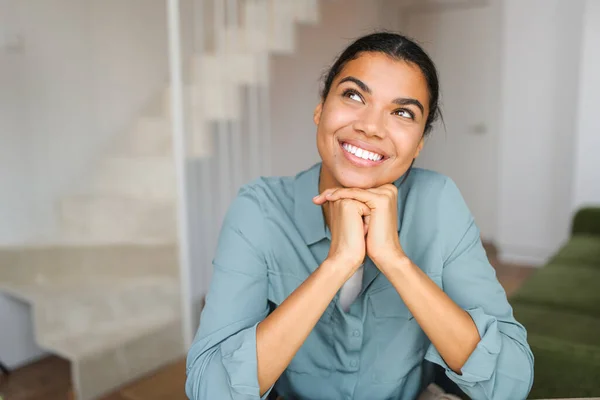 Headshot of young dreaming African American ethnic female with beautiful smile and looking away while sitting against blurred home or office interior — Stockfoto