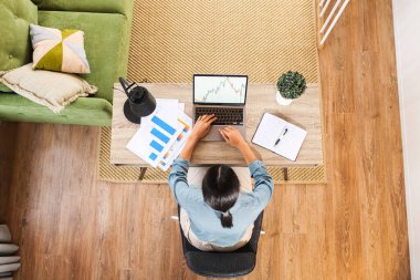 Masada oturan ve dizüstü bilgisayarda yazan melez etnik kadın ya da kadın serbest yazar genel bakış açısına göre not alıyor. Modern ev ofisi