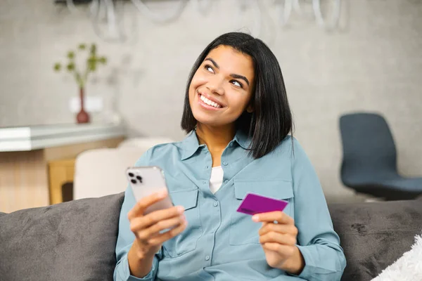 Serene smiling dreamy multiracial woman holding credit card and smartphone, thinking about long awaited purchase. Happy inspired lady is shopping online, paying for travel voucher — стоковое фото