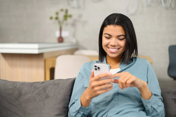 Boldog multiracionális nő ül a kanapén, és időt töltenek online az ő smartphone, SMS-ek, görgetés hírcsatornák, megosztási hozzászólás a szociális hálózatok — Stock Fotó