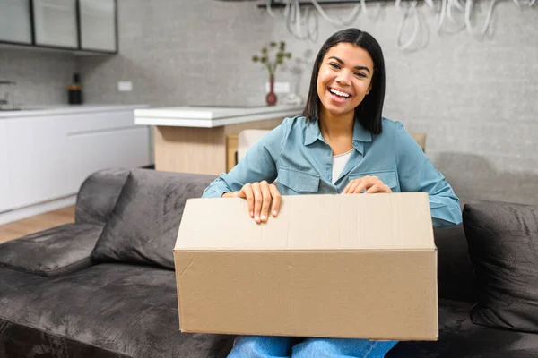Positive young African American woman sitting on the couch with carton box on laps, feeling curious about ordered item from an online store, smiling client satisfied — 图库照片