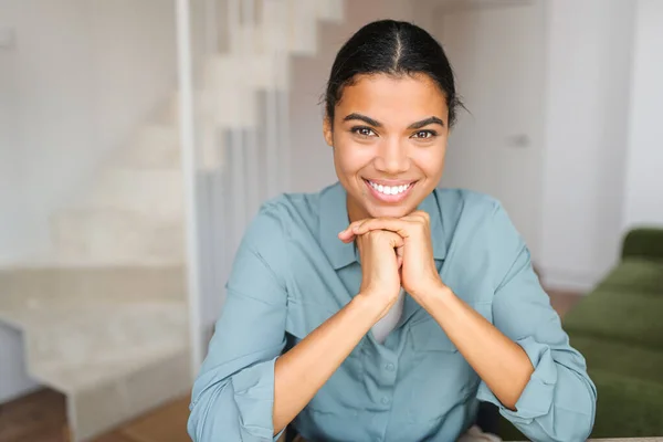 Portrait of charming elegant woman with hands under chin smiling at the camera, sitting in the modern house, wearing shirt — Stockfoto