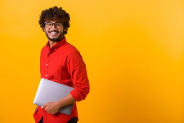 Elinde dizüstü bilgisayarla bir yere giden gülümseyen pozitif Hintli adamın portre görüntüsünü kaldır. Kapalı stüdyo çekimleri turuncu renkte.