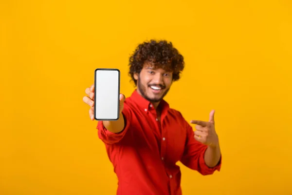 Portrét spokojeného bruneta muže s kudrnatými vlasy ukazuje na mobilní telefon a usmívá se na fotoaparát, doporučuje gadget nebo mobilní aplikace. Vnitřní studio záběr izolované na oranžové — Stock fotografie