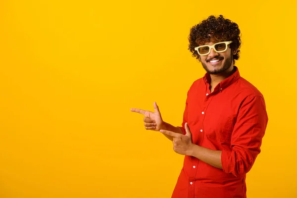 Portrait of pleasant looking bearded man wearing sunglasses pointing finger aside at copy space, showing area for advertisement. Indoor studio shot isolated on orange — Fotografia de Stock