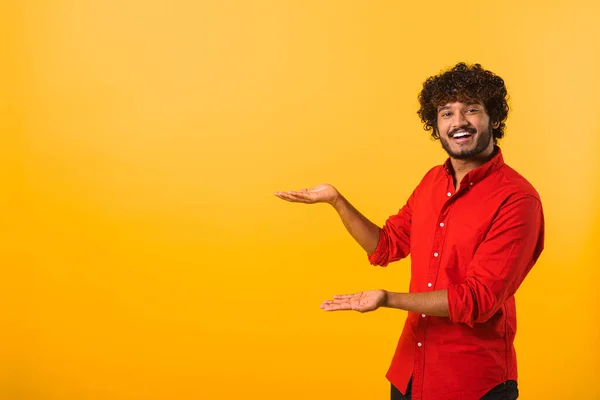 Joyful curly man presenting advertising area on his palm, holding empty place for commercial. Indoor studio shot isolated on orange — 图库照片