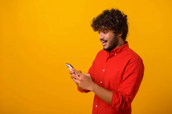 Portrétní pohled na mladého dospělého muže, jak se dívá na mobilní telefon s šokovaným užaslým výrazem s velkýma očima a otevřenými ústy. Vnitřní studio záběr izolované na oranžové — Stock fotografie