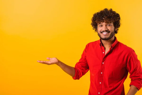 Curly indian man wearing red casual style T-shirt, pointing at copy space on his palm, showing empty place for idea presentation, product advertising. Indoor studio shot isolated — 图库照片