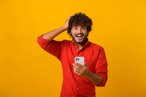 Curly brunette man with beard expressing amazement and holding head with his hands, while using cellphone, with open mouth and shocked big eyes. Indoor studio shot isolated — стоковое фото