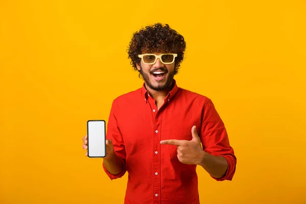 Ευτυχισμένος χαρούμενος χαρούμενος χάντρες δείχνοντας το δάχτυλο στο smartphone με άδεια οθόνη κοιτάζοντας κάμερα με χαμόγελο toothy, ελεύθερος χώρος για adv. Εσωτερική στούντιο πυροβόλησε απομονώνονται σε πορτοκαλί — Φωτογραφία Αρχείου